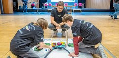 Das Motto des diesjährigen Wettbewerbs: „Roboter in der Landwirtschaft“ Drei Jugendliche arbeiten auf einem Spielfeld am Boden an Teilen eines Roboters.