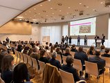 Ein Saal voller sitzender Menschen hört einer Rede zu. 