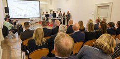 Preisverleihung P-Seminar-Preis Man sieht das Publikum von hinten vor einer großen, weißen Leinwand sitzen. Auf der Bühne stehen mehrere Leute vor einem Rednerpult.