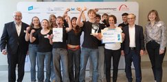 Die Schüler*innen der Rudolf-Steiner-Schule Gröbenzell Jubelnd steht eine Gruppe von Schüler*innen zusammen mit vier Erwachsenen vor einer Leinwand und halten zwei Urkunden in den Händen.