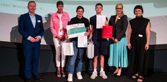 Zwei Jungen stehen mit Urkunden und Geschenken zwischen vier Erwachsenen, drei Frauen und einem Mann, vor einer Leinwand.