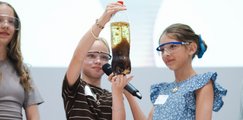 Drei Mädchen führen ihr Experiment, eine Flasche mit einer Mischung aus verschiedenen Flüssigkeiten, vor. 