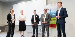Zwei Frauen, ein Mann sowie zwei Schüler stehen vor einer Leinwand nebeneinander und diskutieren miteinander.