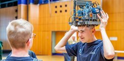 Die 6. Bayerische Robotik-Meisterschaft Ein Jugendlicher hat sich einen Roboter auf den Kopf gesetzt, wobei ihn ein anderer Junge beobachtet.