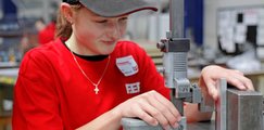 Eine Teilnehmerin in rotem F.EE-T-Shirt arbeitet an einem Messtisch mit einem Höhenmessgerät und überprüft die Maße eines Metallbauteils in einer Werkhalle.