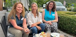 Drei Frauen sitzen zusammen, essen Kuchen und lächeln in die Kamera.