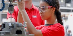 ine Teilnehmerin mit Schutzbrille bedient in einer Werkstatt eine Bohrmaschine. Daneben steht eine weitere Person und unterstützt bei der praktischen Arbeit an der Maschine.
