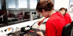 Eine Teilnehmerin arbeitet an einem Elektronik-Arbeitsplatz mit Kabeln, Werkzeugen und Messgeräten. Im Hintergrund befindet sich ein weiterer Arbeitsplatz mit einer zweiten Person.
