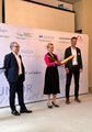 Eine Frau steht zwischen zwei Männern und klappt eine Startklappe zusammen. 