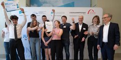 Die Schüler*innen des Willibald-Gluck-Gymnasium München Schüler*innen stehen jubelnd vor einer Leinwand und halten zwei Urkunden in die Höhe. Neben ihnen stehen drei Erwachsene, die Beifall klatschen.