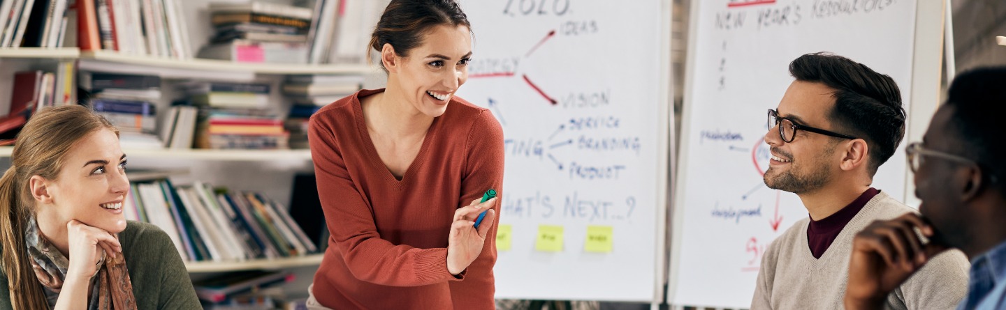 Eine lächelnde Frau steht an einem Tisch mit bunt gestaltetem Flipchart-Papier und kommuniziert mit drei lächelnden und sitzenden Menschen.