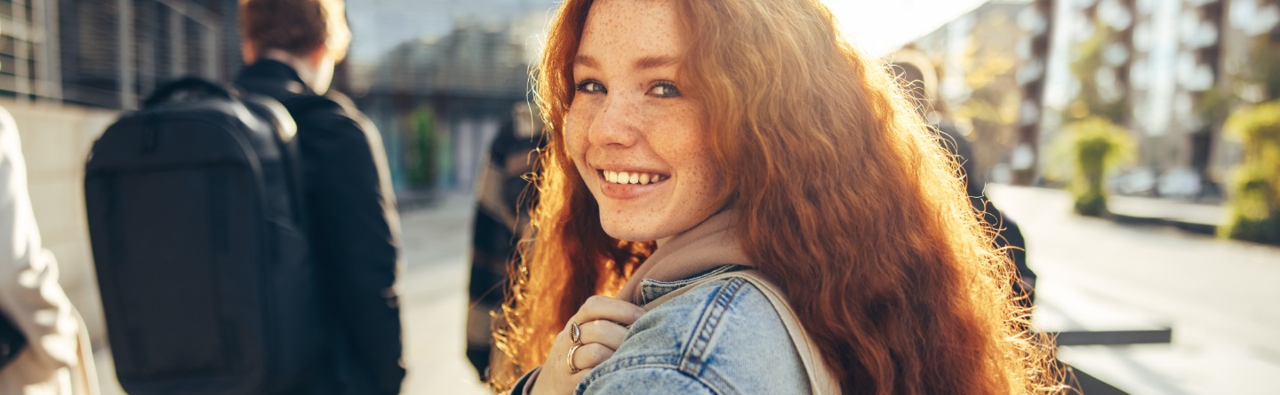 Eine junge Studentin mit langen rötlichen Haaren lächelt in die Kamera. Im Hintergrund sind weitere Studenten von hinten zu sehen.