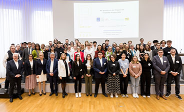 Verleihung des P-Seminar-Preises Eine große Gruppe an Menschen steht vor einer Präsentation auf der Bühne. Es sind junge und ältere Menschen zu sehen, viele sind schick gekleidet.