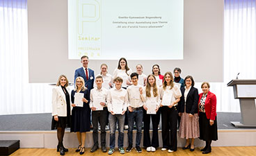 Goethe-Gymnasium Regensburg Auf dem Bild sieht man eine Gruppe Jugendlicher, die lächelnd ihre Urkunden in die Kamera halten. Sie stehen dabei auf einer Bühne.