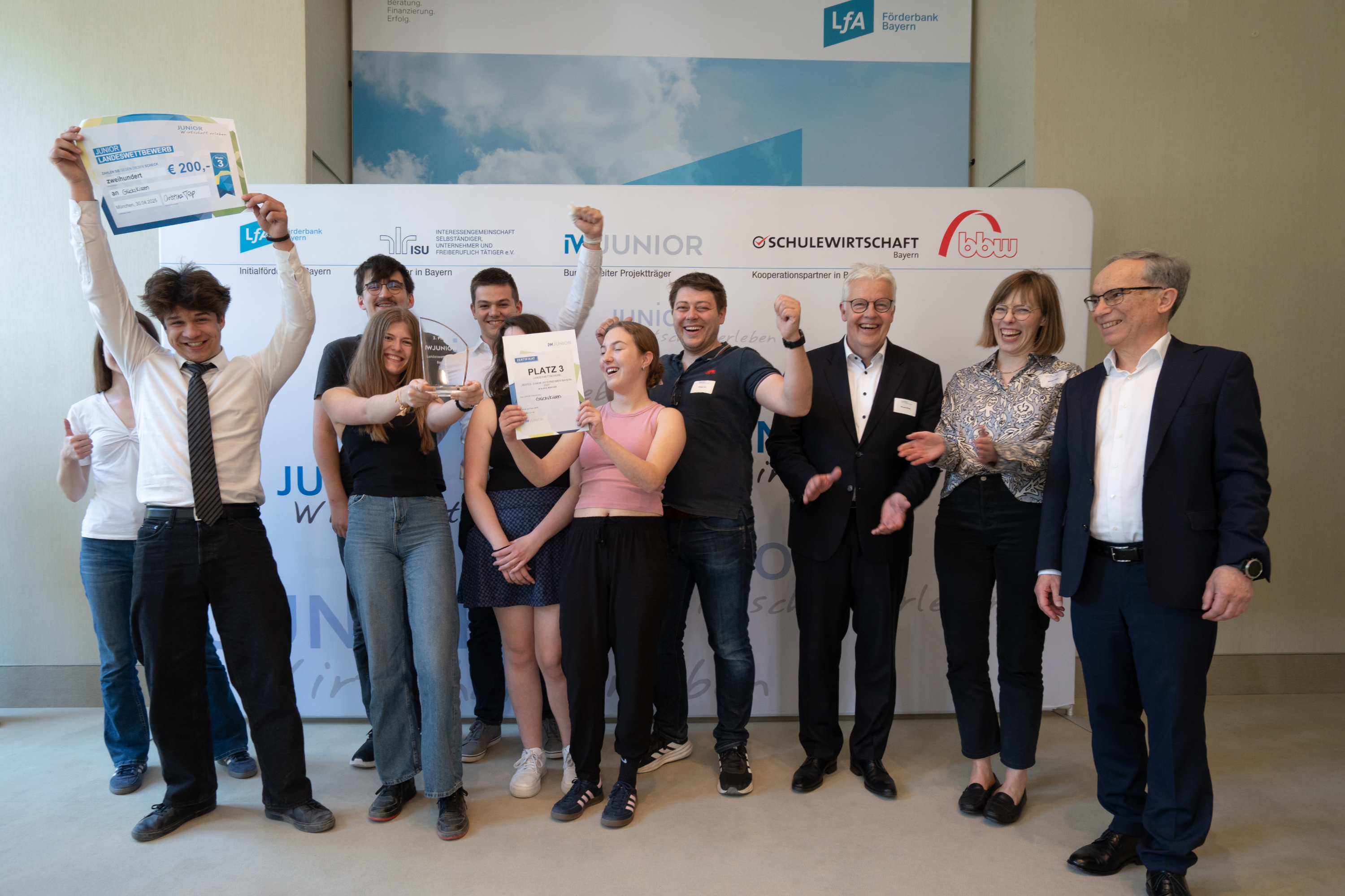 Team „GlücksKissen“ – nachhaltige Sitzkissen für Schulsitze Schüler*innen strecken jubelnd die Hände nach oben und zeigen ihre Urkunden.