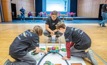 Teilnehmer der Bayerischen Robotik-Meisterschaft Drei Jugendliche arbeiten auf einem Spielfeld am Boden an Teilen eines Roboters.