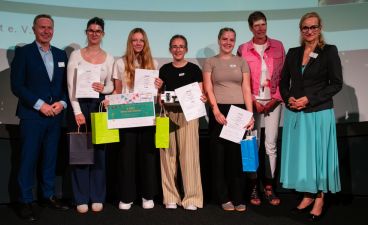 Vier Schülerinnen posieren mit Urkunden und Geschenken für ein Foto. Dabei sind sie von drei Erwachsenen, einem Mann und zwei Frauen, umringt.