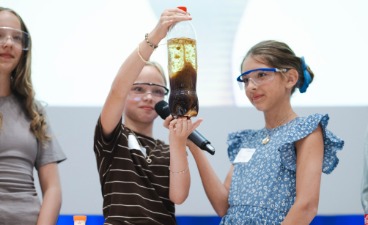 Projekt-Präsentation Drei Mädchen führen ihr Experiment, eine Flasche mit einer Mischung aus verschiedenen Flüssigkeiten, vor.