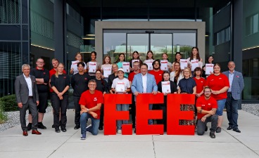 Gruppenfoto vor einem modernen Firmengebäude: Mehrere Jugendliche halten Zertifikate in den Händen und stehen gemeinsam mit Betreuenden hinter großen roten Buchstaben mit der Aufschrift „F.EE“.