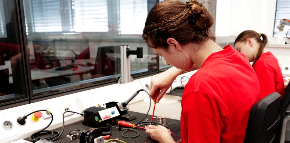 Eine Teilnehmerin arbeitet an einem Elektronik-Arbeitsplatz mit Kabeln, Werkzeugen und Messgeräten. Im Hintergrund befindet sich ein weiterer Arbeitsplatz mit einer zweiten Person.