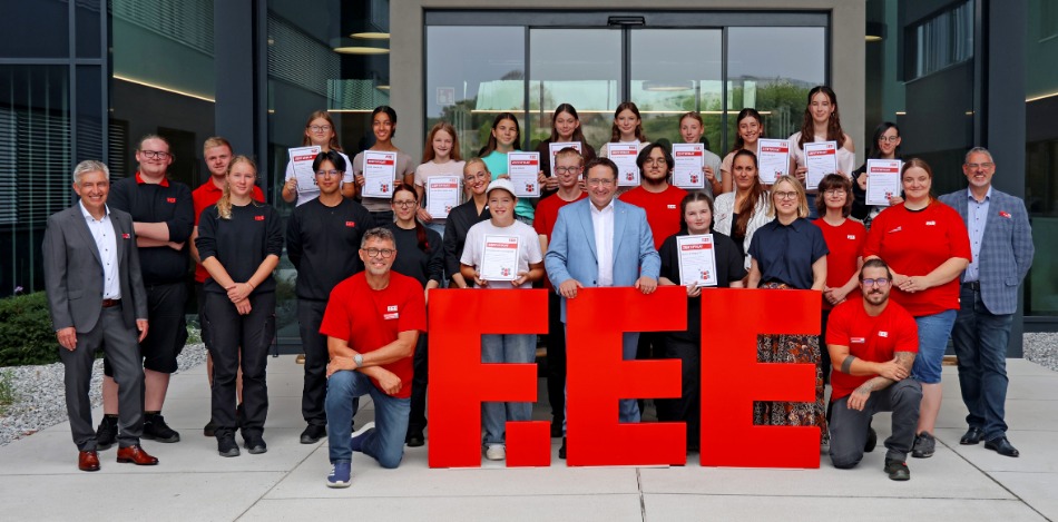 Gruppenfoto vor einem modernen Firmengebäude: Mehrere Jugendliche halten Zertifikate in den Händen und stehen gemeinsam mit Betreuenden hinter großen roten Buchstaben mit der Aufschrift „F.EE“.