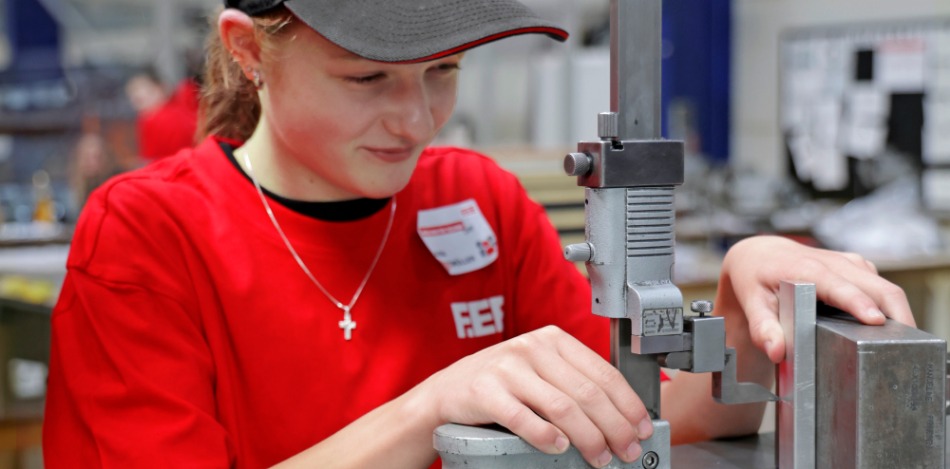 Eine Teilnehmerin in rotem F.EE-T-Shirt arbeitet an einem Messtisch mit einem Höhenmessgerät und überprüft die Maße eines Metallbauteils in einer Werkhalle.
