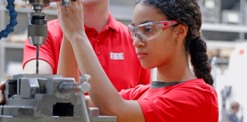 Eine Teilnehmerin mit Schutzbrille bedient in einer Werkstatt eine Bohrmaschine. Daneben steht eine weitere Person und unterstützt bei der praktischen Arbeit an der Maschine.