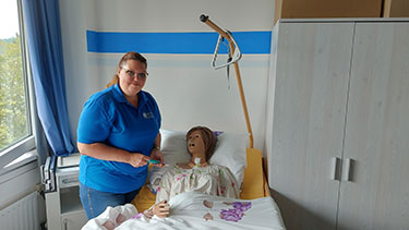 Eine Frau steht neben einem Krankenbett, in dem eine Simulationspuppe aus der Pflege liegt. 