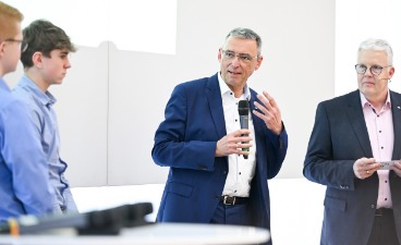 Ein Mann hält eine Rede. Zwei weitere Männer schauen in dabei an.
