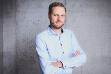 Ein Mann in einem hellblauen Hemd steht mit verschränkten Armen vor einer grauen Wand.