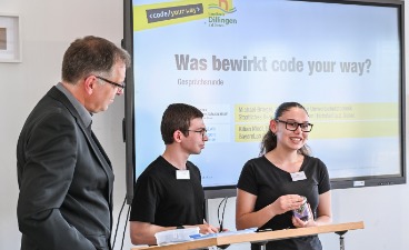 Ein Mann steht mit zwei Schüler*innen an einem Pult. Im Hintergrund sieht man eine Präsentation.