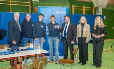 Gruppenbild mit 7 Personen die nebeneinander in einer Turnhalle in die Kamera schauen. Direkt hinter ihnen steht ein Roll-up und vorne links sieht man einen Teil eines Tisches mit Give-Aways drauf