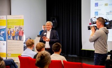 Eine Person hält einen Vortrag vor einem Publikum. Ein Kameramann filmt die Szene. Im Hintergrund sind ein Plakat und eine Leinwand zu sehen.