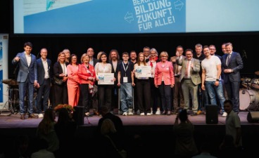 Eine Gruppe von Personen steht auf einer Bühne und posiert für ein Foto. Drei von ihnen halten einen Gewinnscheck für ein Superpraktikum in den Händen. Im Hintergrund ist eine große Leinwand mit dem Text ‚BERUFLICHE BILDUNG ZUKUNFT FÜR ALLE!‘ zu sehen.