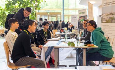 © sprungbrett bayern / Jakob Klinger Eine Gruppe von fünf sechs Menschen sitzt an einem Tisch in einer hellen, modernen Halle. Im Hintergrund sind weitere Personen und Informationsstände zu sehen. Auf dem Tisch liegen Informationsmaterialien und ein Laptop.