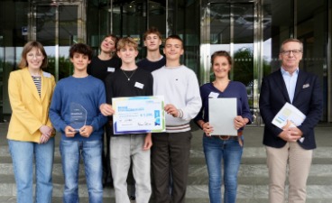 JUNIOR-Bundeswettbewerb Eine Gruppe Schüler*innen posiert auf einer Treppe mit einer Urkunde in der Hand, ein Mann und eine Frau stehen daneben.