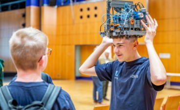 Die 6. Bayerische Robotik-Meisterschaft in Amberg Ein Junge hält einen selbstgebauten Roboter über seinem Kopf und spricht währenddessen mit einem anderen Jungen.