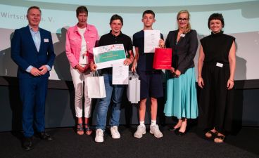 Platz 1 bei Technik-Scouts 2025 Zwei Jungen stehen mit Urkunden und Geschenken zwischen vier Erwachsenen, drei Frauen und einem Mann, vor einer Leinwand.