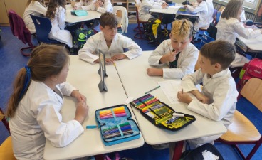 Siegerteam Es funktioniert?!-Auszeichnung 2025: Grundschule Kist Drei Schüler und eine Schülerin sitzen mit weißen Laborkitteln an einem Tisch und besprechen ihre Forscherfrage