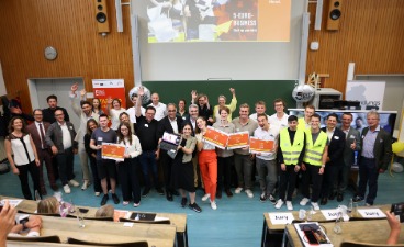 Gruppe von Personen mit Zertifikaten vor einer Leinwand mit dem Schriftzug „5-EURO-BUSINESS“ und Jury-Tischen im Vordergrund.
