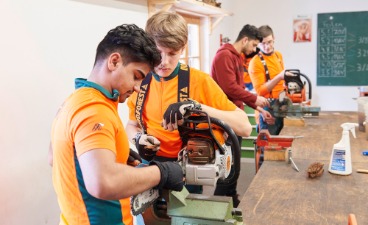 Personen arbeiten in einer Werkstatt an Motorsägen, die in Schraubstöcken eingespannt sind.
