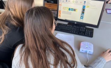 Zwei Personen arbeiten gemeinsam an einem Computer, auf dessen Bildschirm eine grafische Programmieroberfläche zu sehen ist.