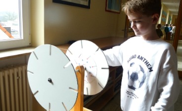 Junge Person dreht an einer Experimentierstation mit zwei weißen Scheiben in einem hellen Schulflur.