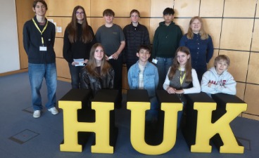 Eine Gruppe von zehn Jugendlichen posiert lächelnd hinter großen, gelben Buchstaben, die das Wort „HUK“ bilden