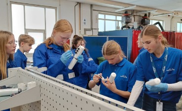 Mehrere weibliche Jugendliche in blauer Arbeitskleidung montieren in einer Werkstatt konzentriert kleine, weiße Bauteile.