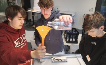 Drei Jugendliche führen in einem Zimmer ein Experiment mit einem Trichter und einer Flüssigkeit durch.