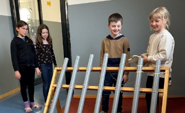 ier Grundschulkinder experimentieren an einer Station der MINIPHÄNOMENTA mit verschieden langen Kunststoffrohren auf einem Holzgestell.