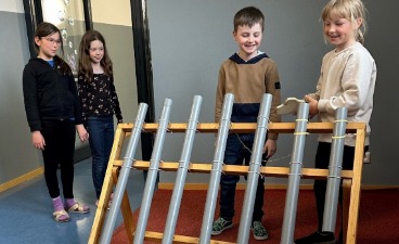 Mehrere Schulkinder stehen vor einer schräg aufgebauten Holzkonstruktion mit parallel angeordneten Metallrohren und beobachten beziehungsweise berühren die Rohre, während sie den Aufbau zum Experimentieren nutzen.