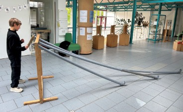Ein Junge steht in einer hellen Schulaula an einer Experimentierstation der MINIPHÄNOMENTA. Die Station besteht aus einem Holzgestell, an dem mehrere lange, graue Kunststoffrohre schräg nach unten verlaufen. Der Schüler hält ein Objekt an die Rohröffnung, um Akustikphänomene zu erforschen. Im Hintergrund ist der großzügige Flurbereich der Schule mit weiteren Lernstationen zu sehen.