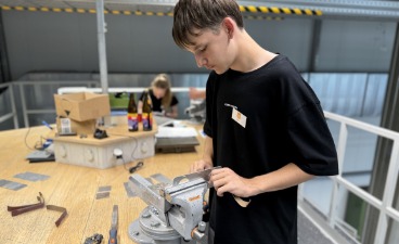 Ein Jugendlicher in einem schwarzen T-Shirt steht in einer Ausbildungswerkstatt an einer Werkbank und bearbeitet konzentriert ein Metallwerkstück, das in einem Schraubstock eingespannt ist. Im Hintergrund ist eine weitere Person an einem Arbeitsplatz zu sehen.